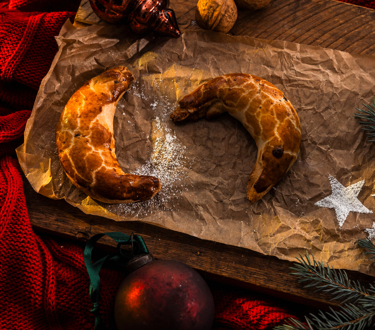 Two crescent-shaped Pressburger Kipferl cookies on crumpled brown paper with Christmas decorations around.