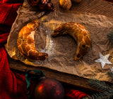 Two crescent-shaped Pressburger Kipferl cookies on crumpled brown paper with Christmas decorations around.