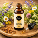 Propolis tincture bottle with liquid in dropper and propolis granules on a wooden plate with flowers in the background.