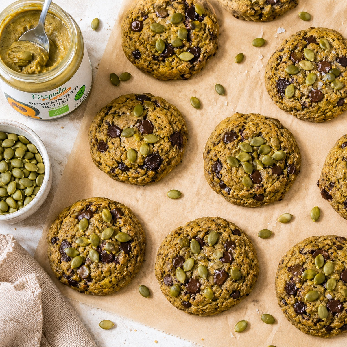 Cookies with pumpkin seeds and chocolate chips on a baking sheet, with a jar of pumpkin seed butter and a bowl of seeds in the background.