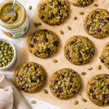 Cookies with pumpkin seeds and chocolate chips on a baking sheet, with a jar of pumpkin seed butter and a bowl of seeds in the background.