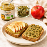 Two slices of bread with pumpkin seed spread, apples, and pumpkin seeds on a plate, with a jar of pumpkin seed butter in the background.