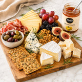 Wooden platter with assorted cheeses, crackers, fruits, and a jar of quince jam on a marble surface.