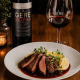 Dinner setting with a bottle of red wine, slices of venison steak in a black pepper jus, and mashed potatoes.