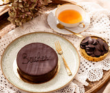 A Sacher Chocolate cake on a plate, accompanied by a cup of tea and chocolate pieces.