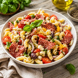 Pasta salad with salami, tomatoes, and basil in a white bowl on a wooden table.
