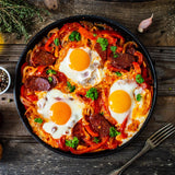 Skillet of shaksuka with eggs, chorizo, and vegetables on a wooden surface