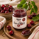 A jar of sour cherry preserve, the jam is on a wooden chopping board surrounded by fresh fruit.