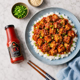 Plate of tofu with spicy ketchup sauce on a bed of rice next to a bottle of Rex Spicy Ketchup on a marble surface.