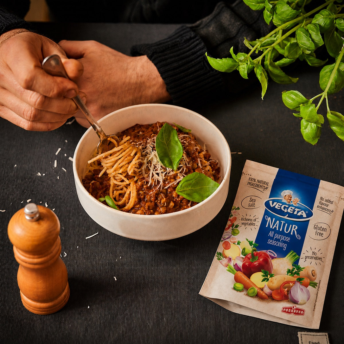 Person about to eat spaghetti with a bowl of pasta, basil leaves, and Vegeta Natur seasoning blend on a dark surface.