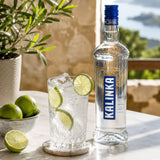 A bottle of Kalinka vodka with a glass of vodka and tonic made with tonic water and lime wedges on a table outdoors.