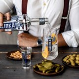 Person pouring Kalinka vodka into a glass with traditional Russian snacks on a wooden table.