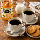 Two cups of coffee with a jar of wildflower honey and a bowl of cookies on a wooden table.